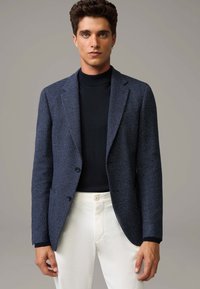 Marineblazer mit strukturiertem Gewebe, zwei Knöpfen und Reverskragen, kombiniert mit einem schwarzen Rollkragenpullover und weißen Hosen.
