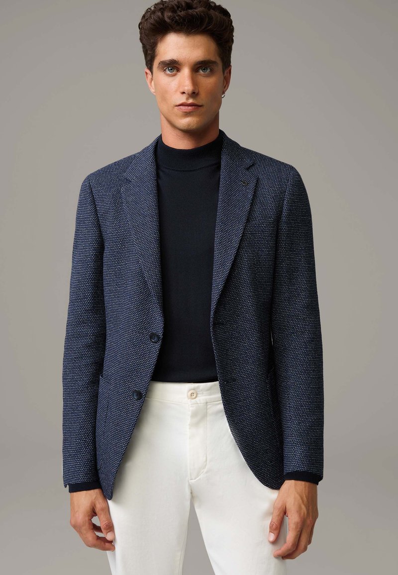 Marineblazer mit strukturiertem Gewebe, zwei Knöpfen und Reverskragen, kombiniert mit einem schwarzen Rollkragenpullover und weißen Hosen.