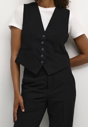 Femme portant un gilet noir boutonné par-dessus un t-shirt blanc à manches courtes et un pantalon noir assorti, posant devant un mur blanc uni.