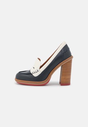 Zapato de tacón de cuero azul marino con un acento blanco y costuras decorativas. Cuenta con un tacón apilado marrón y un detalle en la suela roja.