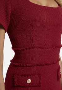 Ein rotes Kleid mit quadratischem Ausschnitt, strukturiertem gewebtem Stoff, fransigen Akzenten an der Taille und zwei Taschenklappen mit goldenen Knopfdetails.