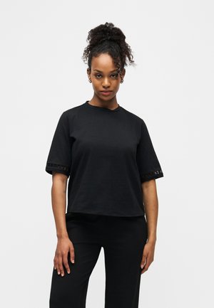 Junge Frau mit lockigem Haar zu einem hohen Dutt gebunden, trägt ein schwarzes Kurzarmoberteil und schwarze Hose, steht vor einem schlichten hellen Hintergrund.