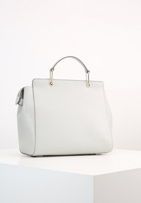 Biała, strukturalna torebka z metalowymi akcentami w kolorze złotym, z pojedynczym uchwytem u góry oraz gładką skórzaną fakturą, wyeksponowana na białej powierzchni.