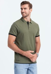 Olivově zelená polo tričko s černými akcenty na límci, krátkými rukávy a zapínáním na tři knoflíky. Bavlněný materiál s logem na lemu.