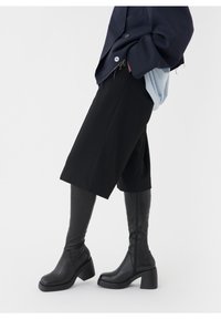 Mustat polvitaihysbootit, joissa on paksu korko, sileä nahkapinta ja sivuvetoketju. Yhdistetty tumman ylisuureen bleiseriin ja mustiin shortseihin.