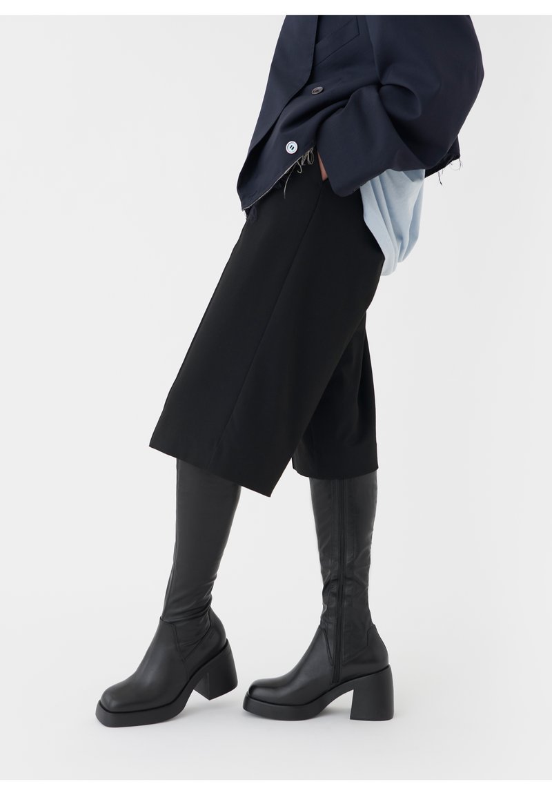 Mustat polvitaihysbootit, joissa on paksu korko, sileä nahkapinta ja sivuvetoketju. Yhdistetty tumman ylisuureen bleiseriin ja mustiin shortseihin.