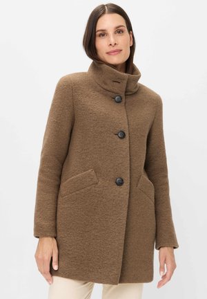 Frau trägt einen braunen Wollmantel mit hohem Kragen und drei schwarzen Knöpfen, steht vor einem einfachen hellen Hintergrund.