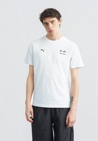 Νεαρός άνδρας με σγουρά μαλλιά, φοράει λευκό T-shirt Puma BMW Motorsport και μαύρο παντελόνι, στέκεται μπροστά σε απλό φωτεινό φόντο.