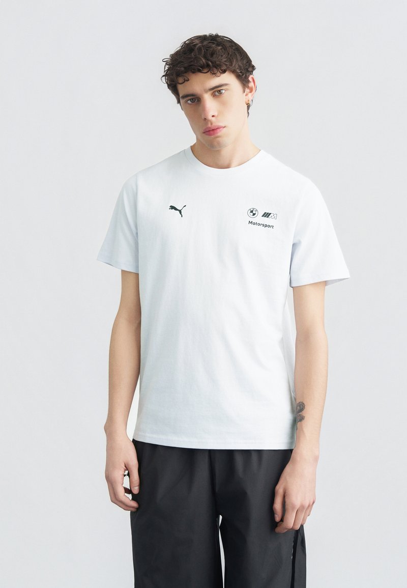 Νεαρός άνδρας με σγουρά μαλλιά, φοράει λευκό T-shirt Puma BMW Motorsport και μαύρο παντελόνι, στέκεται μπροστά σε απλό φωτεινό φόντο.