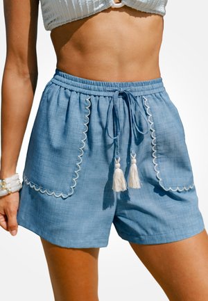 Frau trägt hellblaue Shorts mit Kordelzug, weißen Quasten und gezacktem Saum, kombiniert mit einem weißen strukturierten Crop-Top.