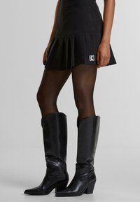 Svart plisserad kjol med logotypdetalj, i kombination med höga svarta läderstövlar med kraftig klack och spetsiga tår, som betonar textur och form.