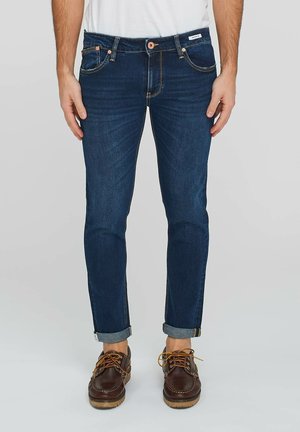 Jeans in denim blu scuro con taglio dritto, leggermente sbiaditi, cuciture in contrasto e risvolti, abbinati a scarpe in pelle marrone.