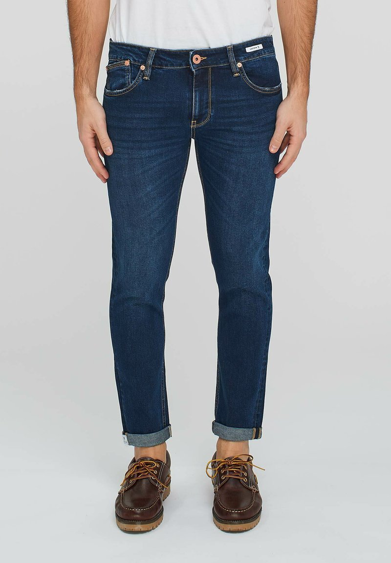 Jeans in denim blu scuro con taglio dritto, leggermente sbiaditi, cuciture in contrasto e risvolti, abbinati a scarpe in pelle marrone.