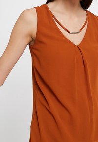 Top senza maniche color ruggine con scollo a V, caratterizzato da una leggera piega e un accento metallico vicino allo scollo. Tessuto morbido e liscio.