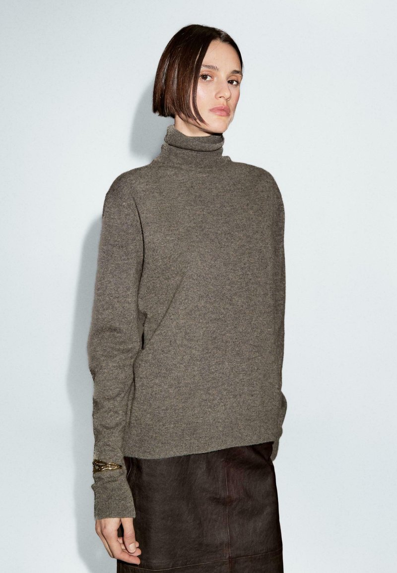 Donna con capelli corti castani che indossa un maglione grigio a collo alto e una gonna in pelle marrone scuro su sfondo bianco uniforme.