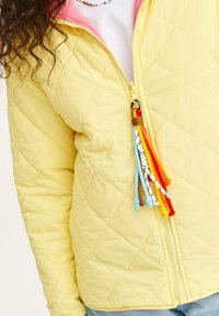 Převlečená žlutá bunda s růžovou podšívkou, se zapínáním na zip a barevnými střapci z provazu. Texturovaná tkanina s diamantovým vzorem.