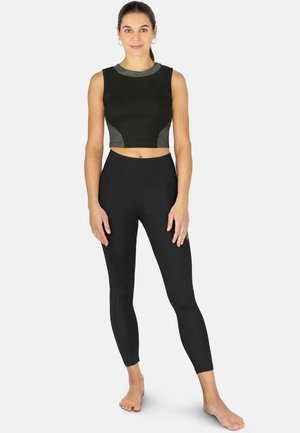 Frau steht barfuß, trägt ein schwarzes ärmelloses Crop-Top und hoch taillierte schwarze Leggings vor einem schlichten hellen Hintergrund.