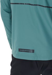 Türkisfarbene, wasserabweisende Jacke mit schwarzem reflektierendem Streifen im oberen Rückenbereich und "ENDURANCE"-Label nahe dem unteren Saum.