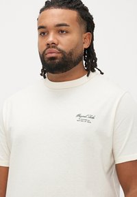 Kremfarvet kortærmet t-shirt i bomuldsblanding med ribstrikket rund hals. Har grøn broderet branding på venstre brystområde.