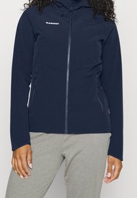 Navy softshell jacket made of flexible material, featuring a zippered front, two side pockets, and a fitted hood. Logo on the chest.