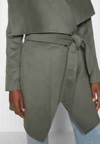 Cappotto avvolgente verde oliva con cintura, orlo asimmetrico, maniche lunghe e texture morbida, caratterizzato da un dettaglio a nodo in vita.