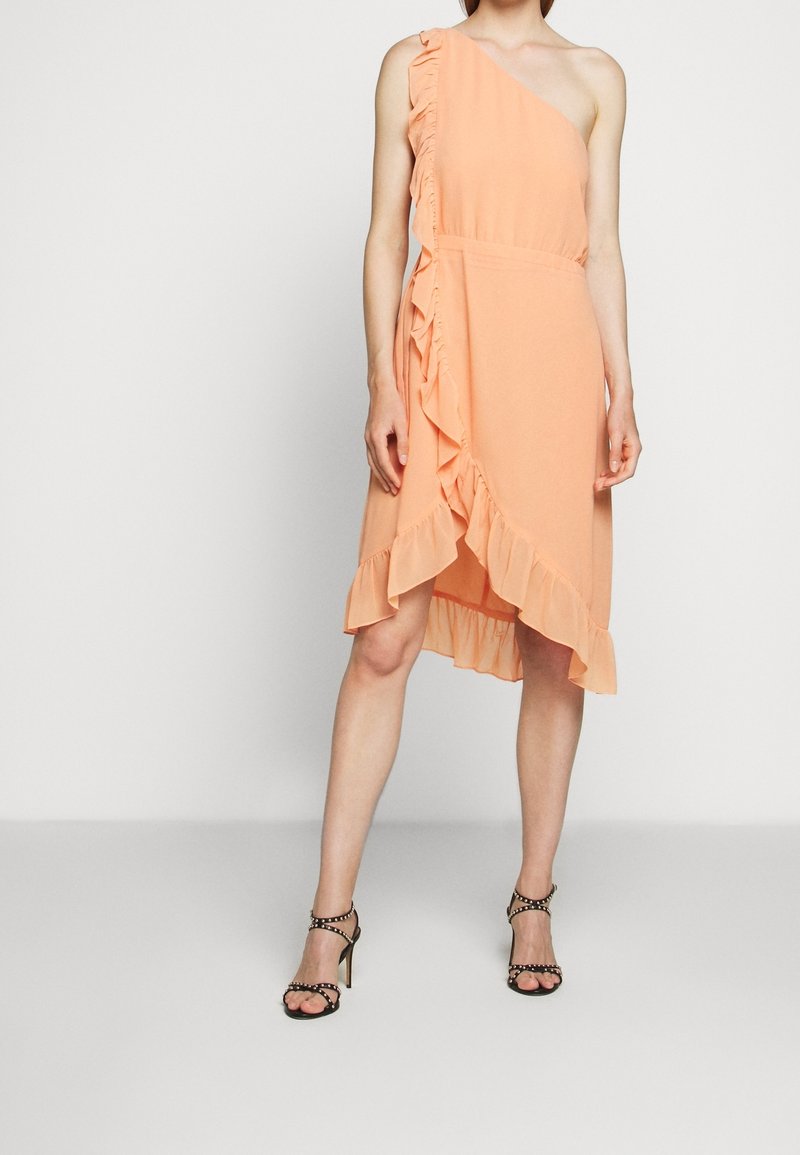 Robe pêche à une épaule avec des volants sur le côté, présentant un ourlet asymétrique. Le modèle porte des talons noirs à brides avec des clous.