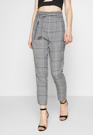 Frau trägt hoch taillierte schwarz-weiße karierte Hose mit Stoffgürtel und schwarzen Knöchelriemen-Absätzen, sichtbares kurz geschnittenes schwarzes Oberteil.