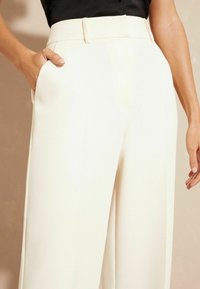 Witte, wijde broek met een gladde textuur, voorzien van zijzakken en een subtiele tailleband. De stof lijkt lichtgewicht en op maat gemaakt.