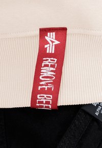 Beige ribfluweel stof met een rood label waarop "REMOVE BEFORE" staat in witte letters. Daaronder is zwarte stof zichtbaar met een contrasterende textuur.