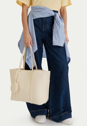 Persona che indossa jeans larghi blu scuro, scarpe da ginnastica bianche, una camicia a righe blu legata in vita e tiene una grande borsa a spalla beige.