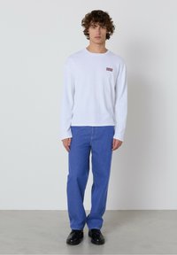 Bijela košulja s dugim rukavima i malim crvenim logotipom s lijeve strane na prsima, uparena s plavim trapericama i crnim cipelama. Jednostavan dizajn, ležeran kroj.