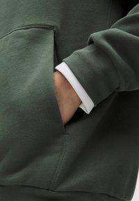 Mano en el bolsillo frontal de un sudadera verde oliva, con un puño de camisa blanca visible debajo de la manga.