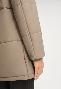 Beżowa ocieplana kurtka z pikowanym wzorem na rękawie, elastycznym mankietem i czerwonym akcentem metki. Miękka, gładka tkanina o prostym kroju.