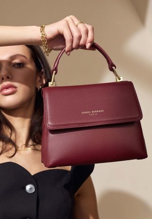 Sac à main en cuir bordeaux avec une forme structurée, une petite poignée, des accents en métal doré et un rabat à l'avant. Nom de la marque embossé à l'avant.