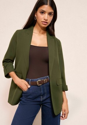 Olivovozelený sakó s upraveným strihom a výstrihom do "V", s vyhrnutými rukávmi a bočnými vreckami, kombinované s hnedým topom a džínsami.