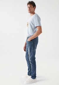 T-shirt azzurro chiaro con un design grafico, abbinato a jeans slim-fit blu medio. Il modello indossa sneakers bianche. Sfondo minimalista.