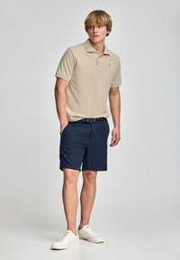 Beżowa koszulka polo z krótkimi rękawami, granatowe szorty i białe sneakersy. Koszulka ma kołnierzyk i małe logo na klatce piersiowej.
