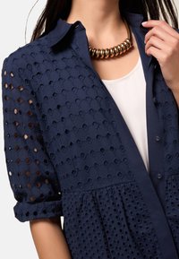 Camicia blu navy con tessuto a nido d'ape, chiusura con bottoni e maniche arrotolate. Indossata sopra un top bianco con una collana dorata e arrotondata.