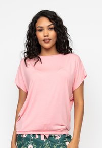 Różowa koszulka z krótkim rękawem, wykonana z lekkiego materiału, o luźnym kroju i okrągłym dekolcie. Połączona ze wzorzystymi zielonymi spodniami.