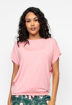 Różowa koszulka z krótkim rękawem, wykonana z lekkiego materiału, o luźnym kroju i okrągłym dekolcie. Połączona ze wzorzystymi zielonymi spodniami.