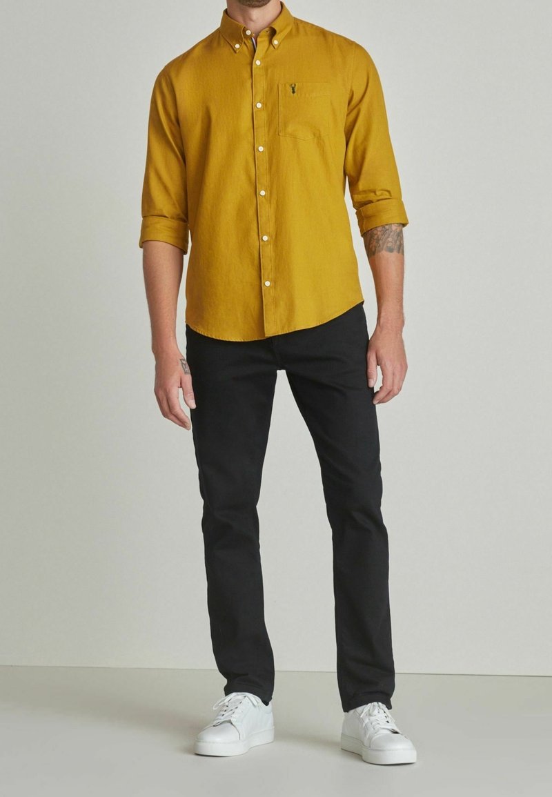 Homme portant une chemise moutarde à boutons avec poche, pantalon noir et baskets blanches, debout devant un fond clair uni.