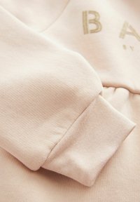 Primo piano di un hoodie rosa chiaro, realizzato in un morbido misto cotone con cuciture visibili e polsini a coste. Dettaglio con la scritta dorata "BA".