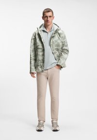 Ελαφρύ, μοτίβο μπουφάν σε πράσινο και μπεζ, συνδυασμένο με ανοιχτό μπλε πόλο, μπεζ παντελόνι και γκρι αθλητικά παπούτσια με μαύρες λεπτομέρειες.