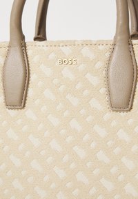 Beige teksturēta rokassoma ar gludām pelēkbrūnām rokturiem. Iezīmēts ar smalku reljefu rakstu un zeltītu "BOSS" logo centrā.