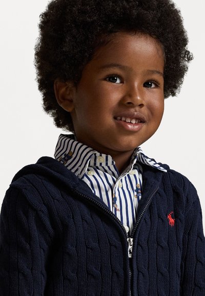 Ναυτική φούτερ ζακέτα με πλέξη καγκουρό και κόκκινο λογότυπο, φορεμένη πάνω από λευκό και μπλε καρό πουκάμισο με μικρά σχέδια.