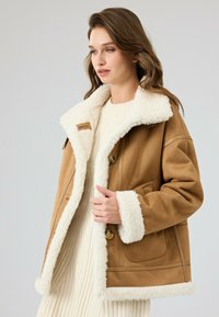 Bruine suede jas met een witte nepbont kraag en manchetten, voorzien van knopen, zakken en een relaxte pasvorm. Gedragen over een crèmekleurige geribbelde trui.