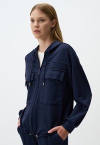 Veste à capuche zippée marine en tissu doux, dotée de deux poches poitrine, d'un ourlet avec cordon de serrage et d'accents dorés sur la fermeture éclair et le tirette.