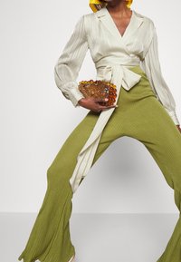 Blouse crème plissée avec un design portefeuille, associée à un pantalon large vert olive texturé. Accessoirisé d'une pochette ambre à motifs.