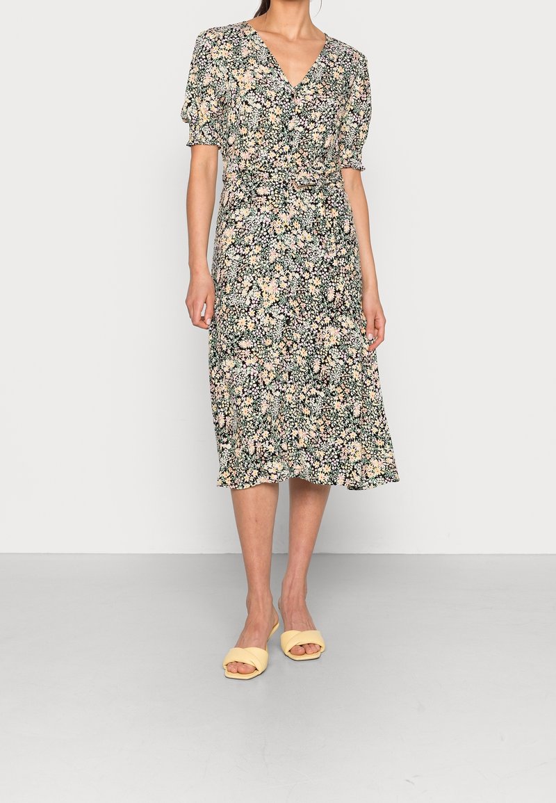 Femme portant une robe midi fleurie avec des manches courtes bouffantes et un col en V, associée à des sandales plates jaunes à bout ouvert sur un fond uni.