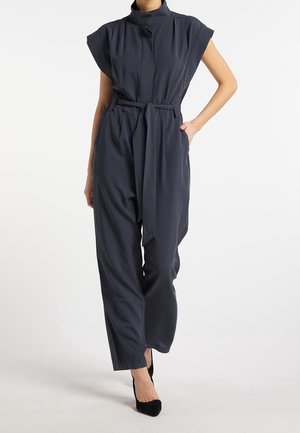 Vrouw in een mouwloze donkerblauwe jumpsuit met riem en wijde pijpen, met zwarte puntige hakken, staand tegen een witte achtergrond.